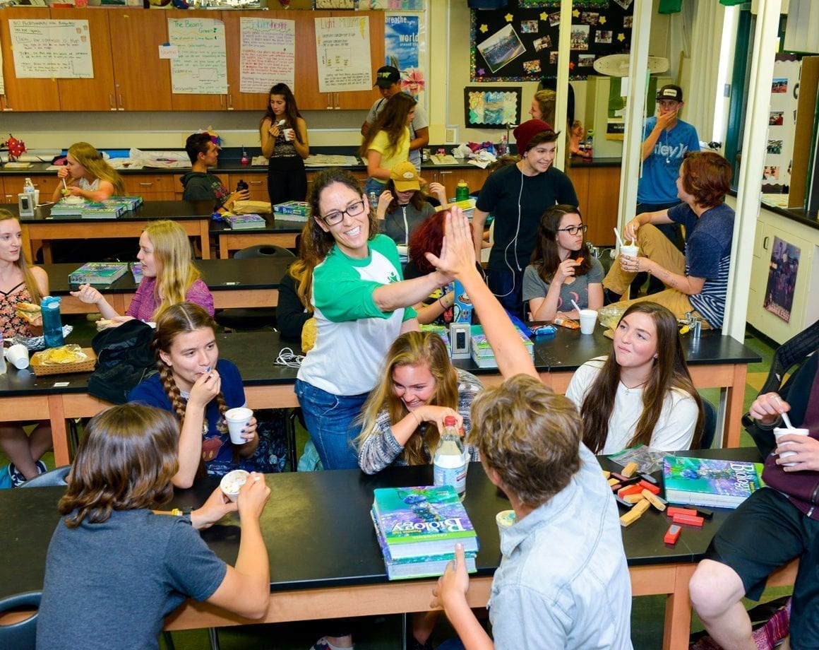 Woman in green and white shirt gives a high five to a student in a classroom surrounded by students at large desks