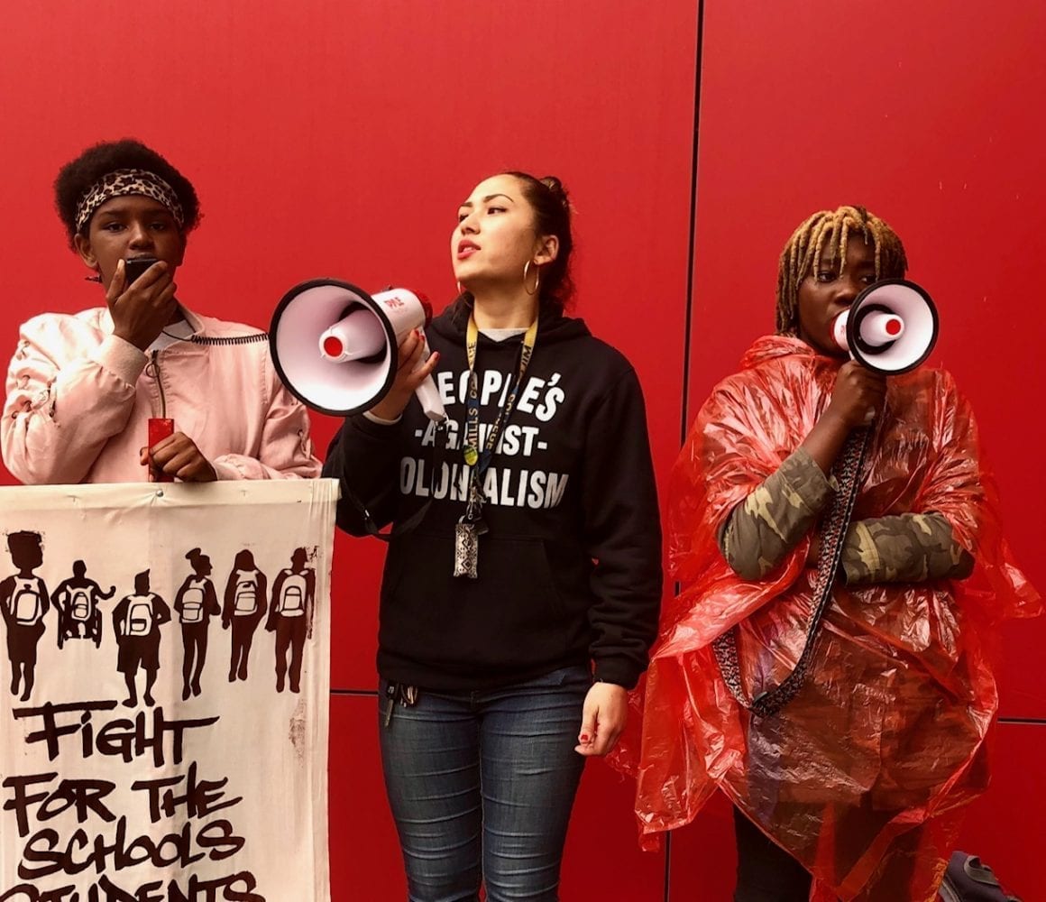 Three women stand in front of red wall holding signs and bullhorns