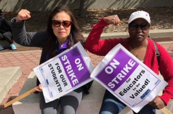 Educators outside sitting down holding strike signs with their solidarity fist pump