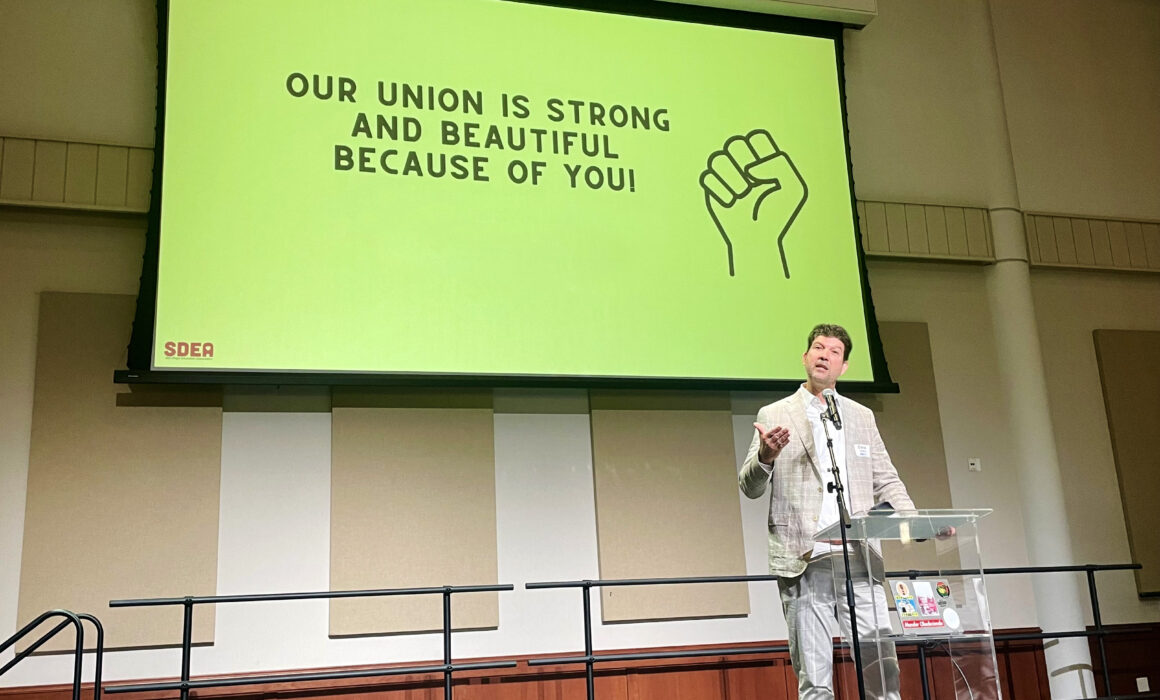 CTA President David Goldberg speaks on stage in front of a green sign that reads "Our Union is Strong and Beautiful Because of You."